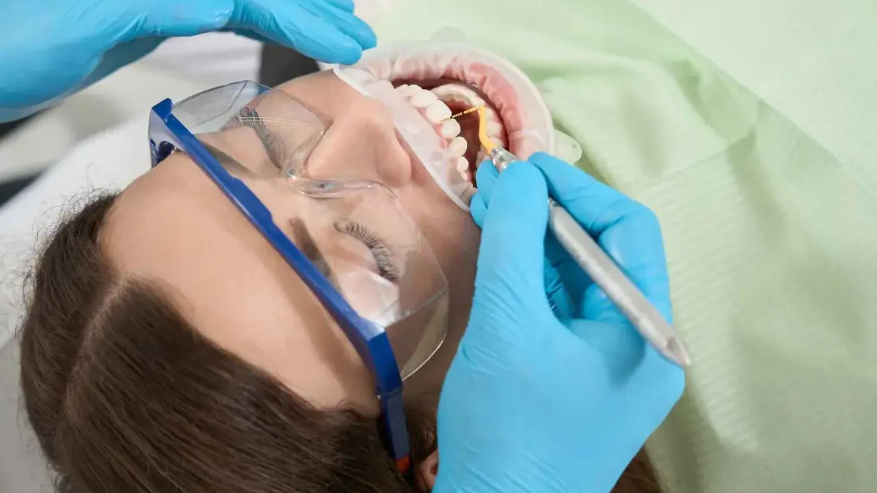 Dentysta wykonuje zabieg higienizacji jamy ustnej. Pacjentka ma założone okulary ochronne i koferdam.
