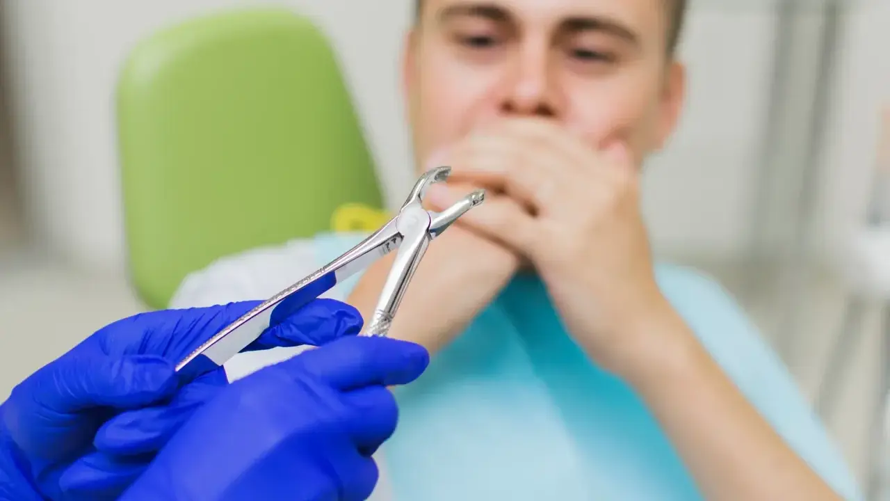 Dentysta w niebieskich rękawiczkach trzyma kleszcze. Pacjent zakrywa usta. Czy po wyrwaniu zęba można wychodzić na dwór?