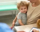 Chłopiec z kręconymi włosami siedzi na kolanach mamy w gabinecie lekarskim. Rozmawiają z lekarzem o jego stanie, być może o cherubizmie.