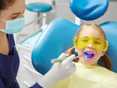 Dziecko w okularach ochronnych u dentysty. Lekarz utwardza lak przy użyciu lampy. Zwykle lakowanie zębów skutki uboczne są minimalne.