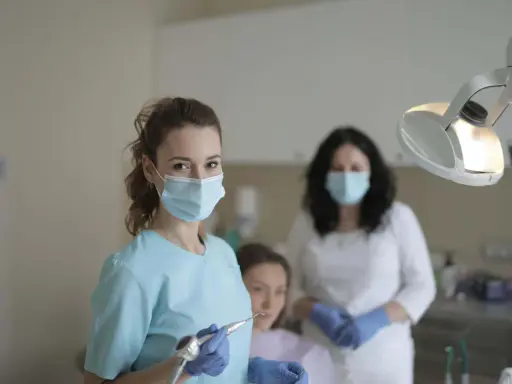 Uśmiechnięta studentka stomatologii w masce i rękawiczkach trzyma wiertło dentystyczne. W tle inne osoby w gabinecie.