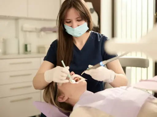 Dentystka w masce i rękawiczkach wykonuje zabieg u pacjentki w gabinecie stomatologicznym.
