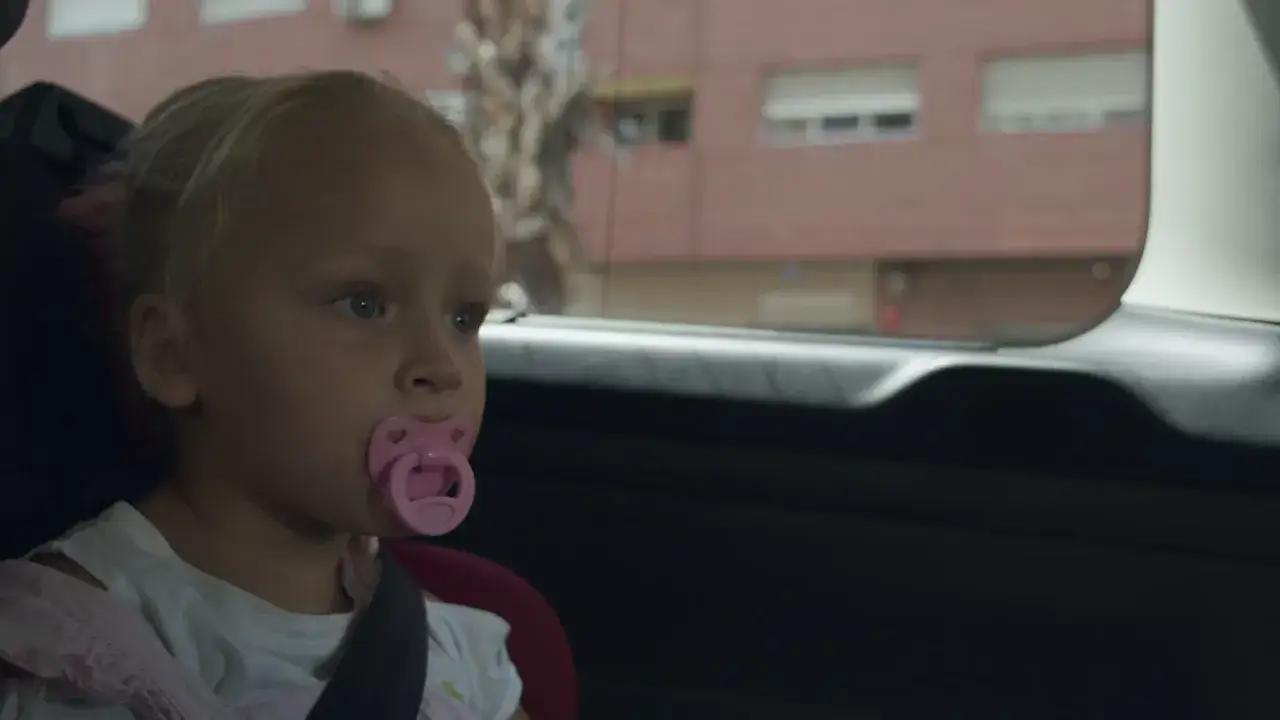 Mała dziewczynka z gryzem otwartym u 2 latka, siedzi w foteliku samochodowym z różowym smoczkiem w buzi.
