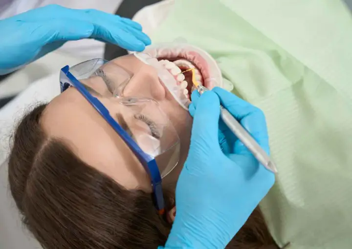 Dentysta wykonuje zabieg higienizacji jamy ustnej. Pacjentka ma założone okulary ochronne i koferdam.