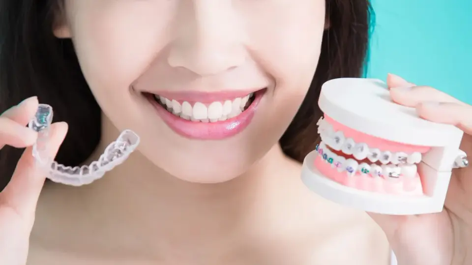 Uśmiechnięta kobieta prezentuje przezroczyste nakładki Invisalign i model szczęki z kolorowym aparatem ortodontycznym.